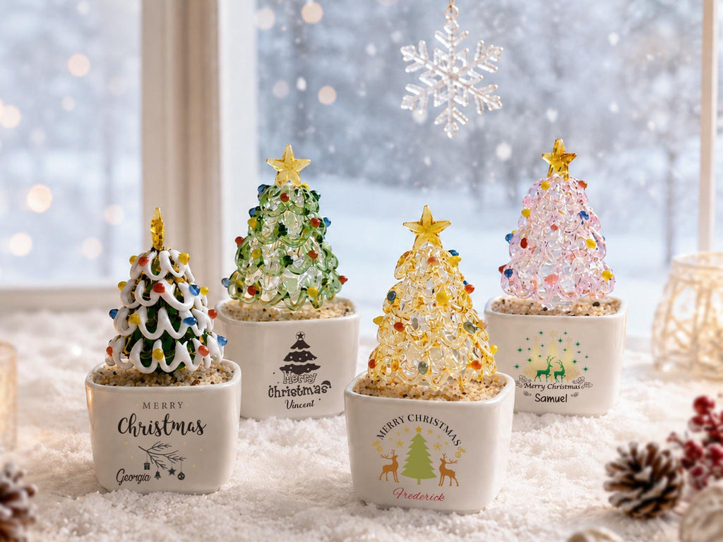 May include: Four miniature glass Christmas trees in white square ceramic pots. Each tree features a gold star topper and colorful glass beads. The pots are printed with Merry Christmas text and names including Georgia, Vincent, Frederick, and Samuel.