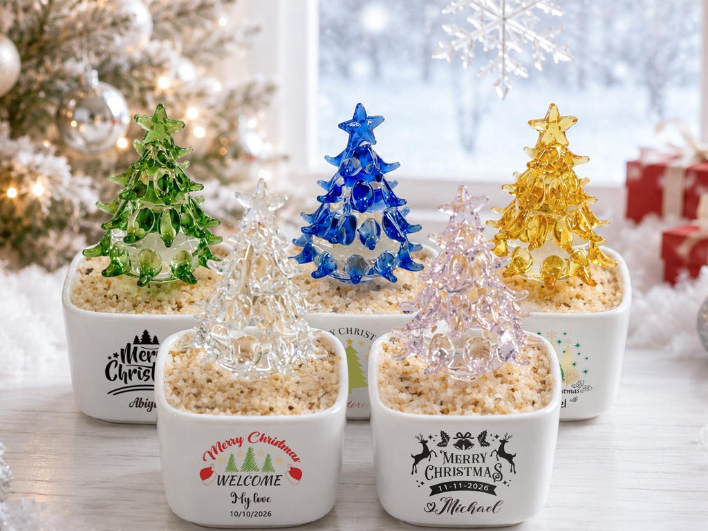 May include: Five small glass Christmas trees in various colors including green, clear, blue, pink, and yellow, each set in a white square ceramic planter filled with tan pebbles. Each planter features black or colored text and holiday graphics.