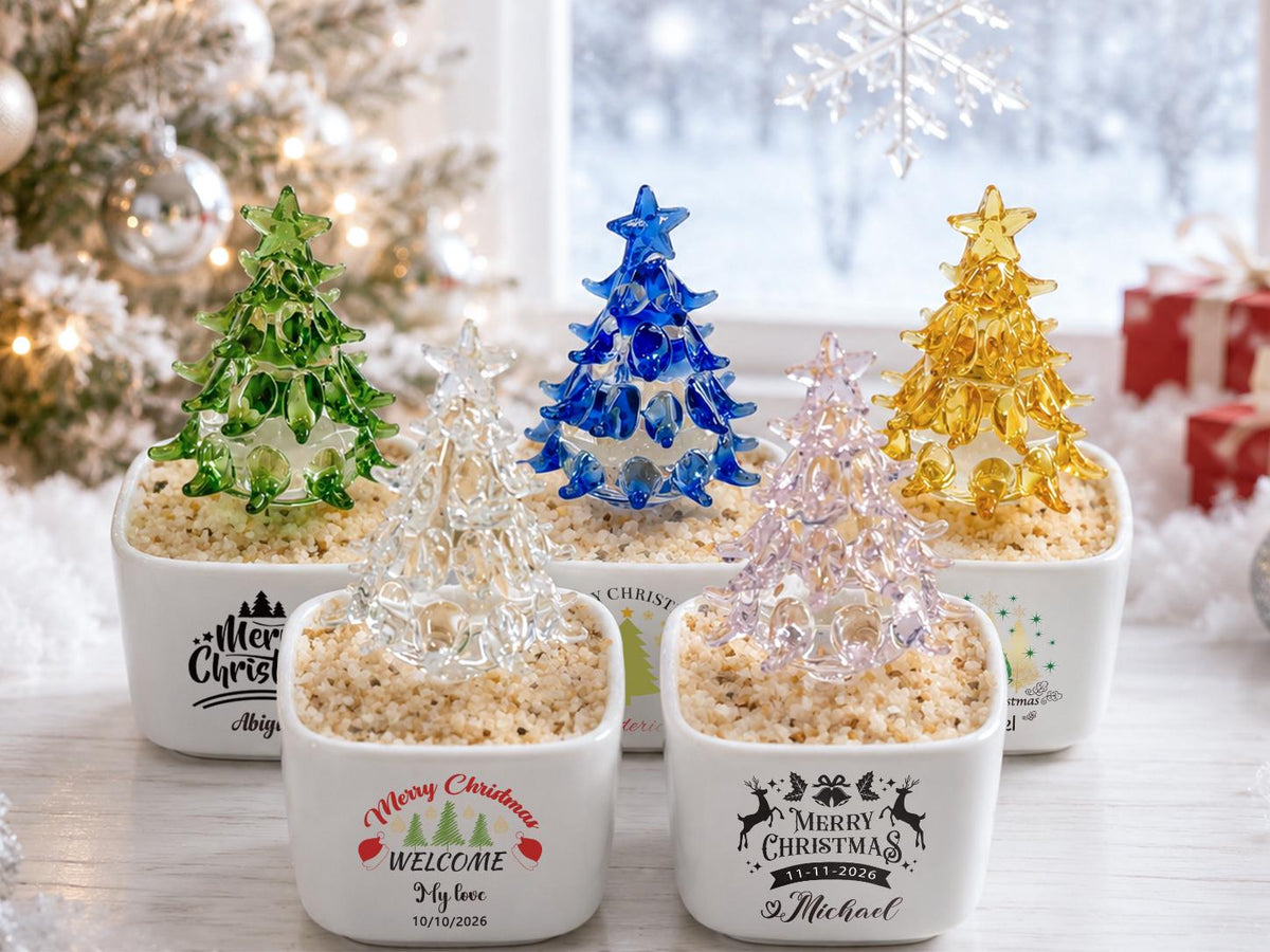 May include: Five small glass Christmas trees in various colors including green, clear, blue, pink, and yellow, each set in a white square ceramic planter filled with tan pebbles. Each planter features black or colored text and holiday graphics.