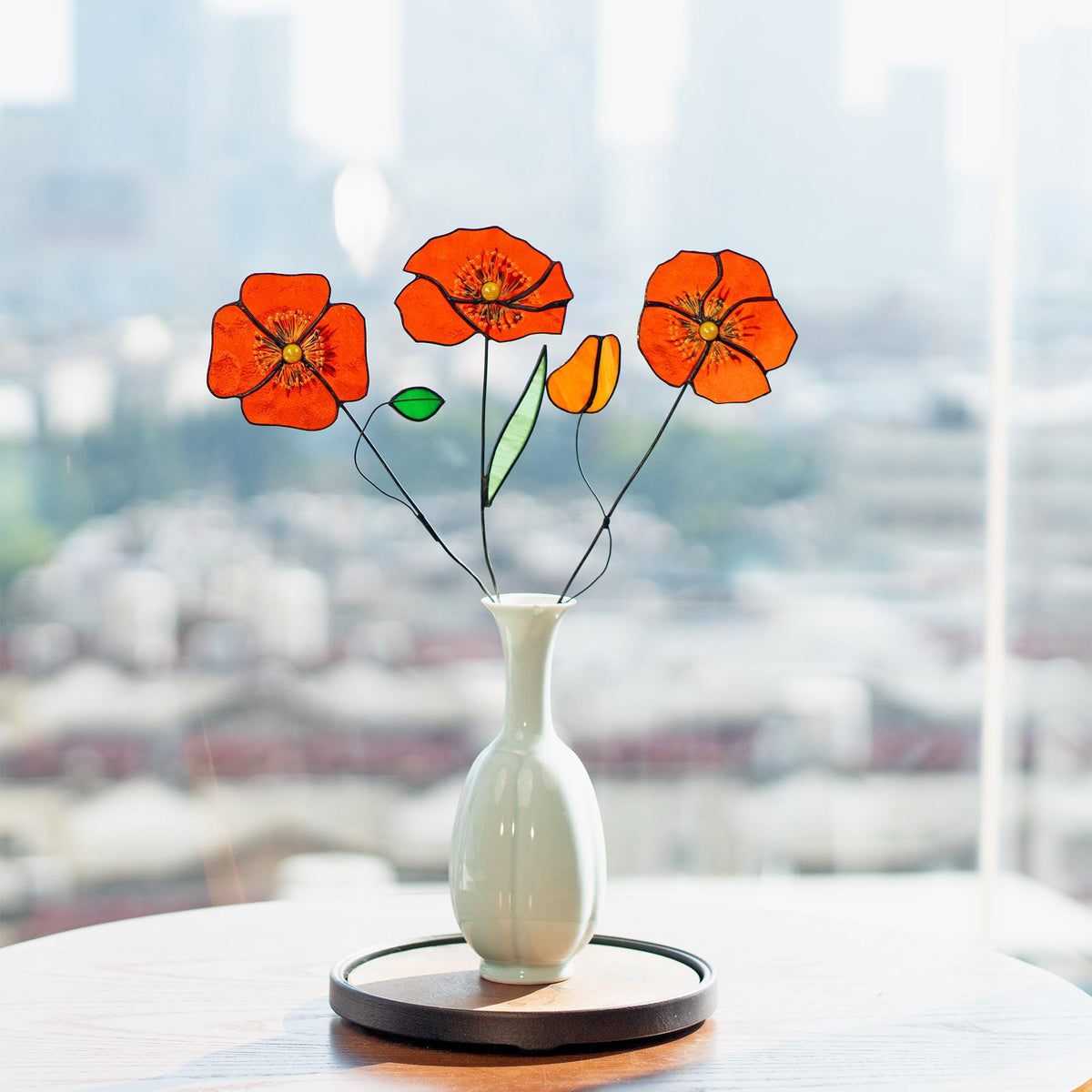 May include: A white ceramic vase holds three orange and yellow stained glass poppy flowers with black stems and a green leaf. The vase sits on a wooden tray, with a blurred cityscape in the background.