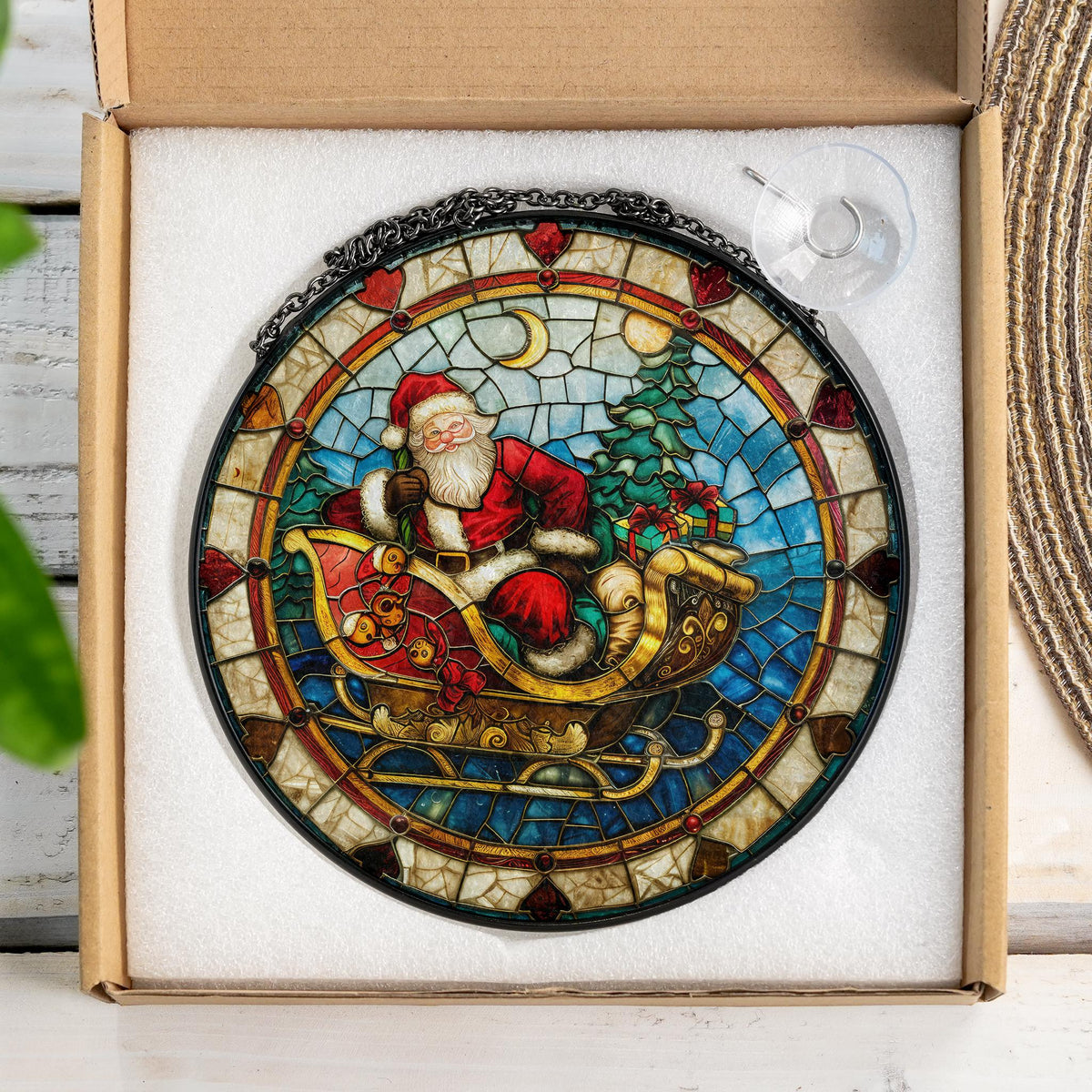 May include: A stained glass window hanging ornament featuring Santa Claus riding in a sleigh with presents. The ornament is round and has a black metal frame. The background is a mosaic of blue, green, and red.