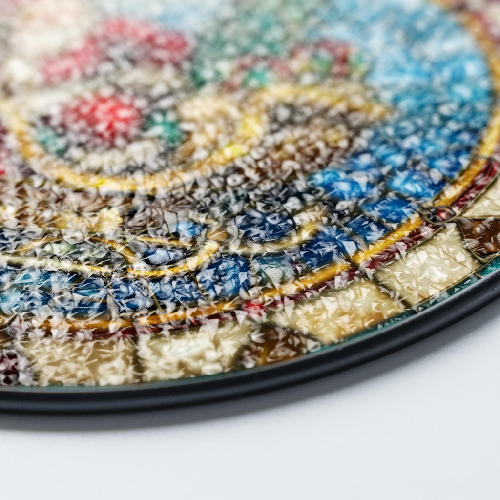 May include: Close-up of a round, colorful mosaic tile with a black rim. The tile has a textured, cracked glass surface.
