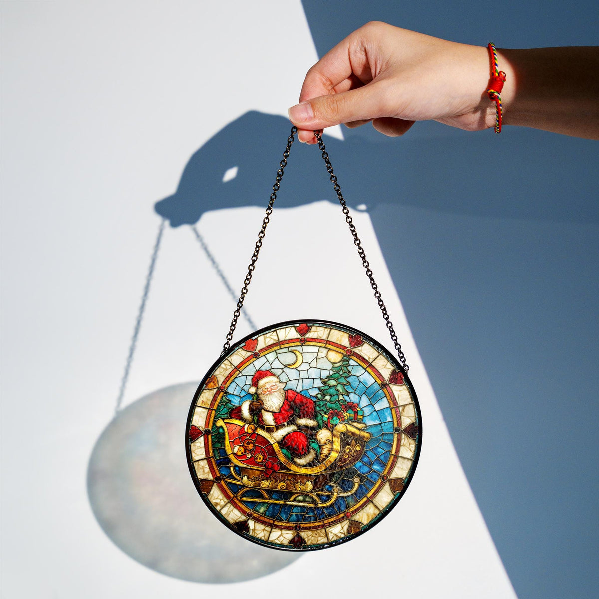 May include: A round stained glass window hanging ornament featuring a depiction of Santa Claus in a sleigh pulled by reindeer. The ornament has a mosaic-like design with red, green, blue, and gold accents.