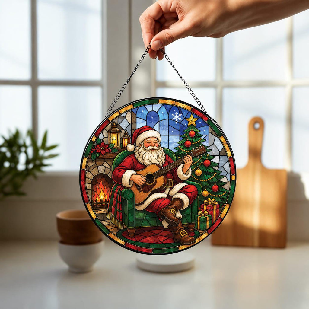 May include: A circular stained glass suncatcher featuring Santa Claus sitting in a green armchair, playing an acoustic guitar. The scene includes a decorated Christmas tree, a fireplace with a glowing fire, and wrapped gifts, all framed in black.