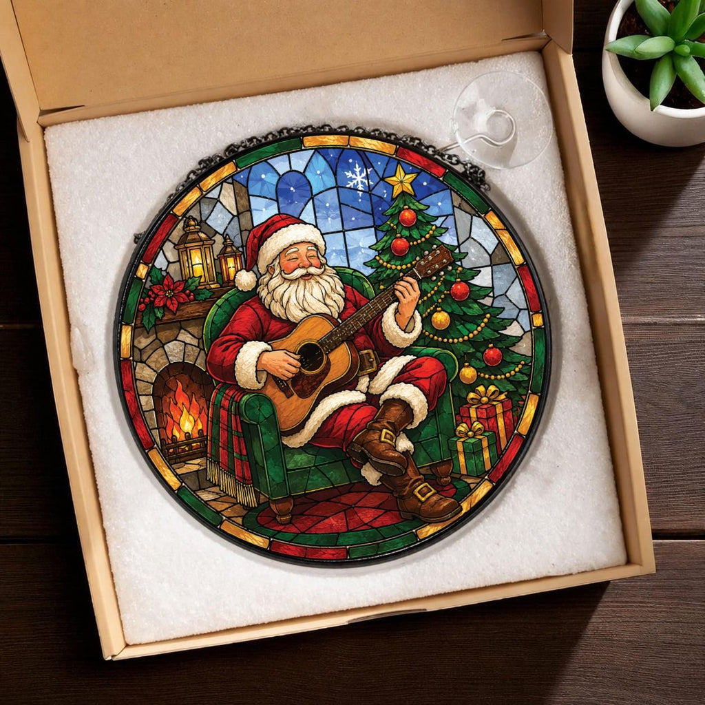May include: A circular stained glass suncatcher featuring Santa Claus sitting in a green armchair playing an acoustic guitar. The scene includes a decorated Christmas tree, a fireplace with a fire, and wrapped gifts, all framed in a dark metal border.