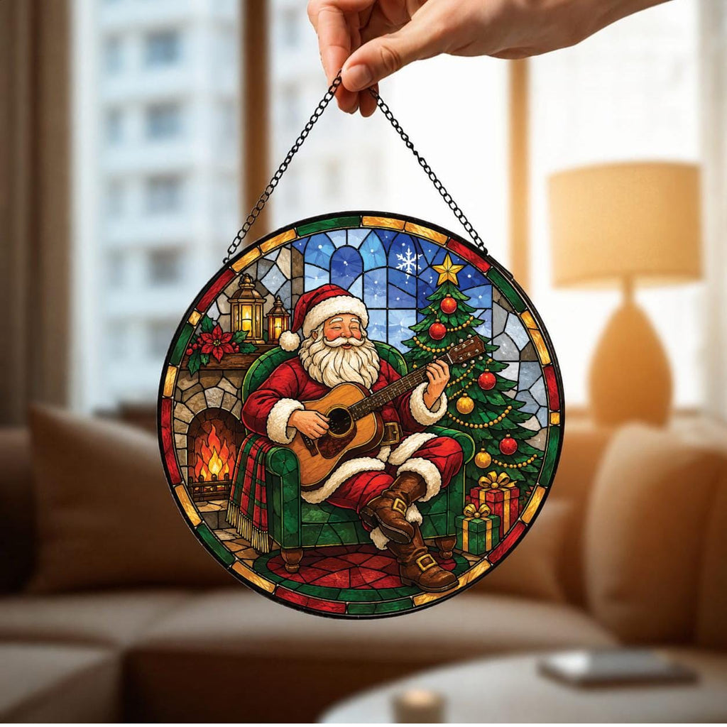 May include: A circular stained glass suncatcher featuring Santa Claus sitting in a green armchair, playing an acoustic guitar. The scene includes a decorated Christmas tree, a fireplace with a lit fire, and wrapped gifts, all framed by a black chain.