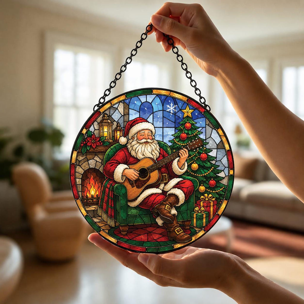 May include: A circular stained glass suncatcher featuring Santa Claus sitting in a green armchair playing an acoustic guitar. The scene includes a decorated Christmas tree, a fireplace with a glowing fire, and wrapped gifts, all framed by a black chain.
