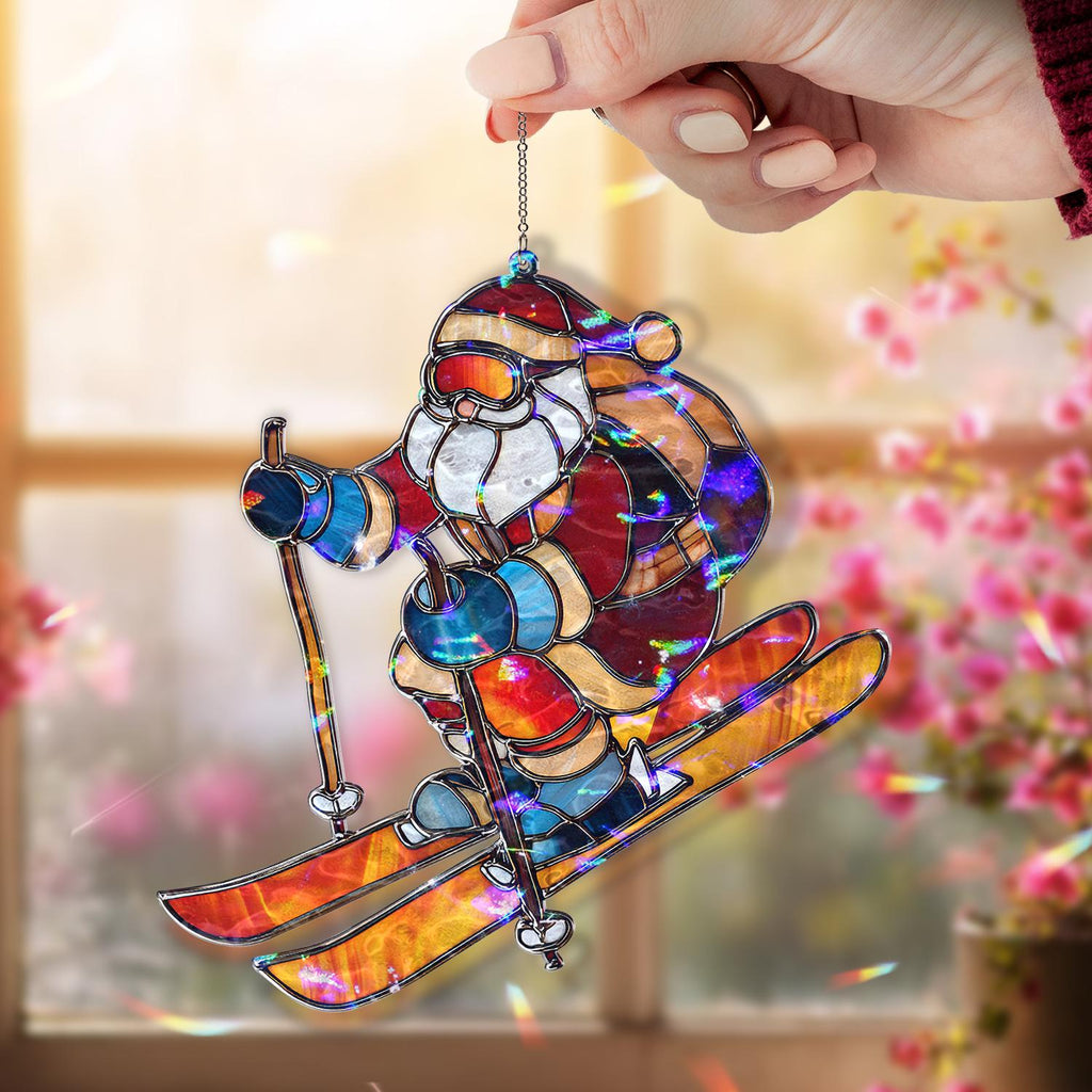 May include: Stained glass ornament of Santa Claus skiing, held by a hand. Santa is wearing a red and white suit, goggles, and a backpack. The skis are orange and yellow. The ornament is hanging from a silver chain.