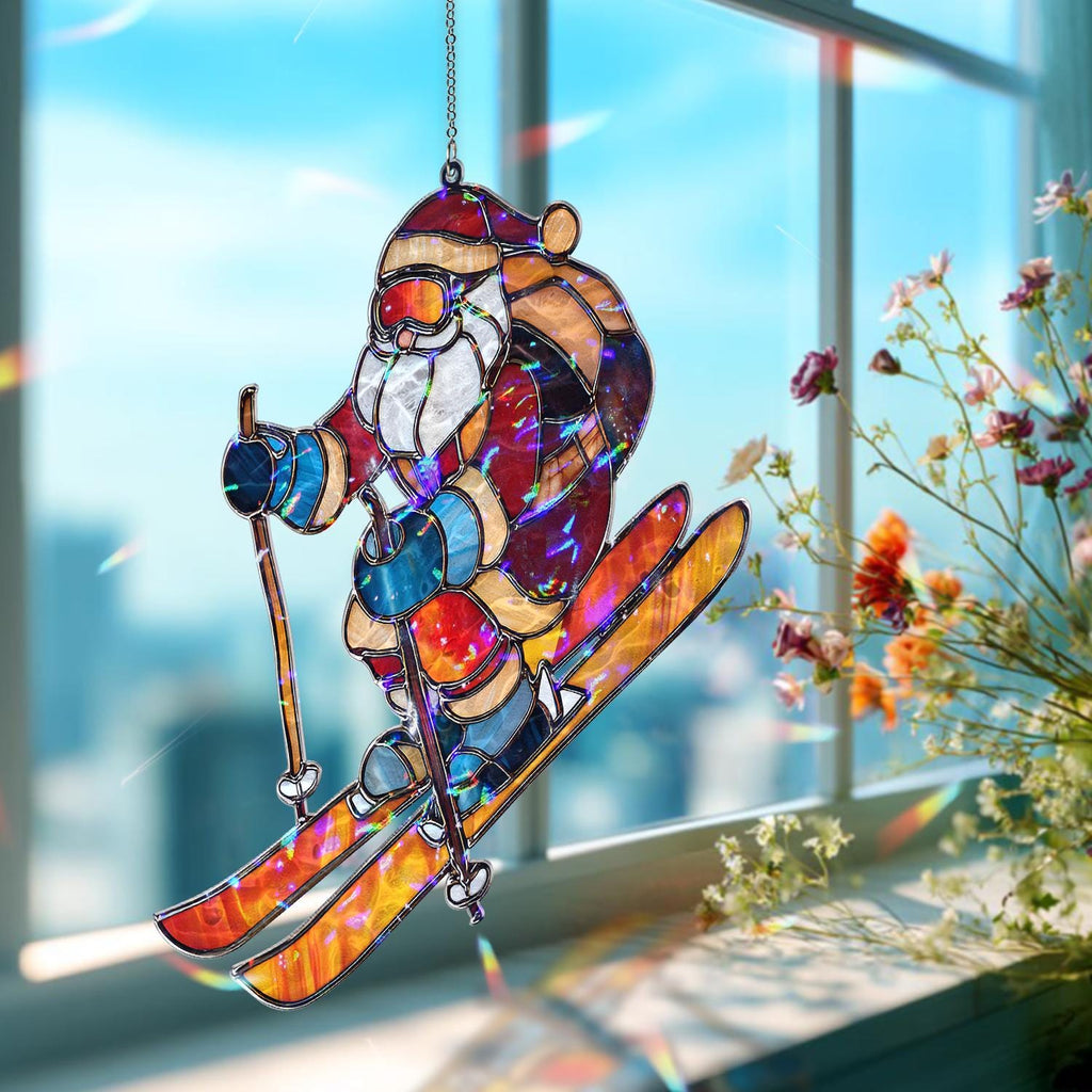 May include: A stained glass Christmas ornament depicting Santa Claus skiing. The ornament features Santa in a red suit, goggles, and a white beard, skiing down a slope. The skis and poles are orange and yellow. The ornament hangs in front of a window.