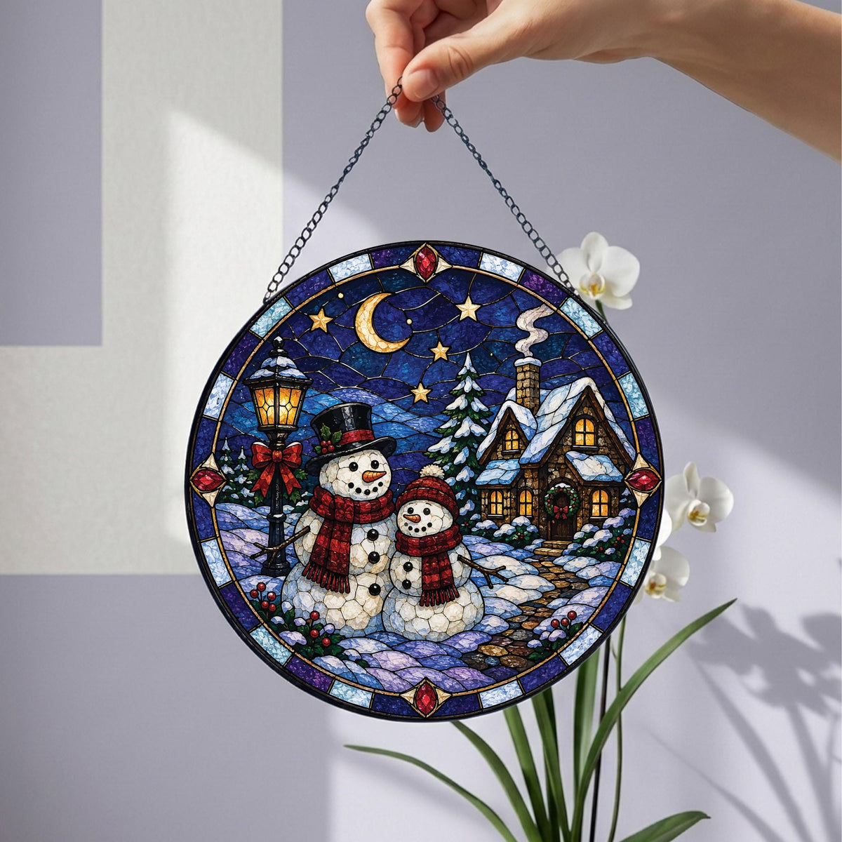 May include: A circular stained glass suncatcher featuring a winter scene with two snowmen, a snow-covered cottage, and a lamppost under a crescent moon and stars. The design is framed by a dark blue border with red accents and hangs from a metal chain.