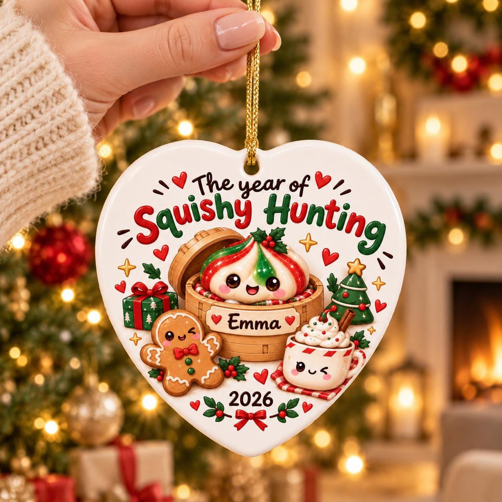 May include: A heart-shaped white ceramic ornament featuring the text The year of Squishy Hunting and the name Emma. The design includes a cartoon dumpling in a bamboo steamer, a gingerbread person, a mug of cocoa, a gift, and the year 2026.