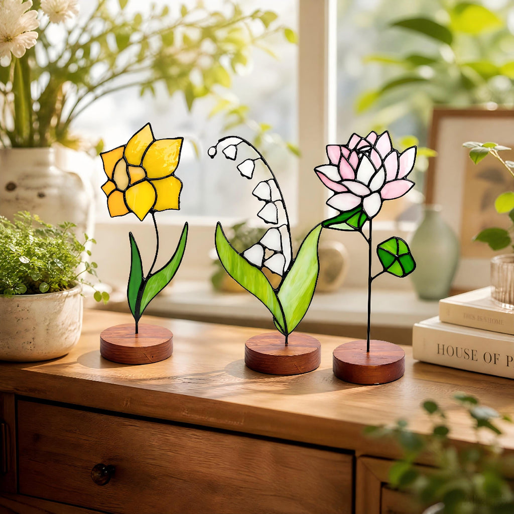 May include: Three stained glass flower sculptures: a yellow daffodil, white lily of the valley, and pink lotus. Each flower is mounted on a small, round wooden base. The flowers are displayed on a wooden surface.