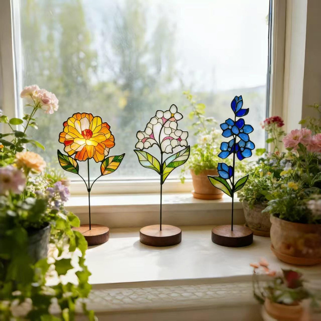 May include: Three stained glass flower sculptures in yellow, white, and blue, each with a black stem and wooden base. The flowers are displayed on a windowsill with potted plants and a window in the background.