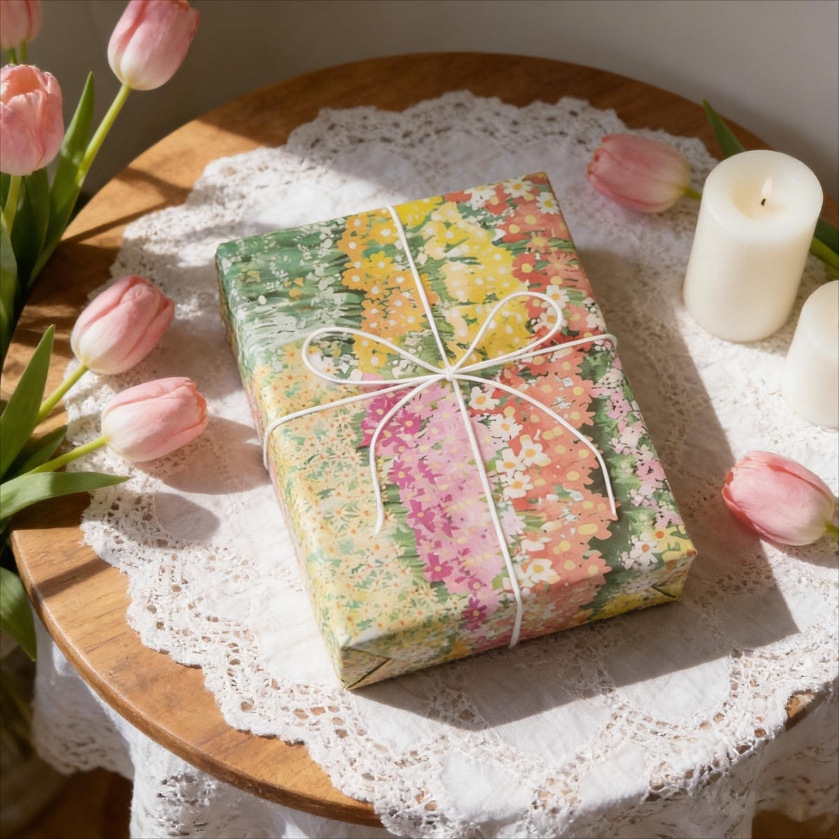 May include: A wrapped gift box with floral patterned paper in shades of yellow, pink, and green, tied with white twine. The box sits on a wooden table with a white lace doily, surrounded by pink tulips and a lit candle.
