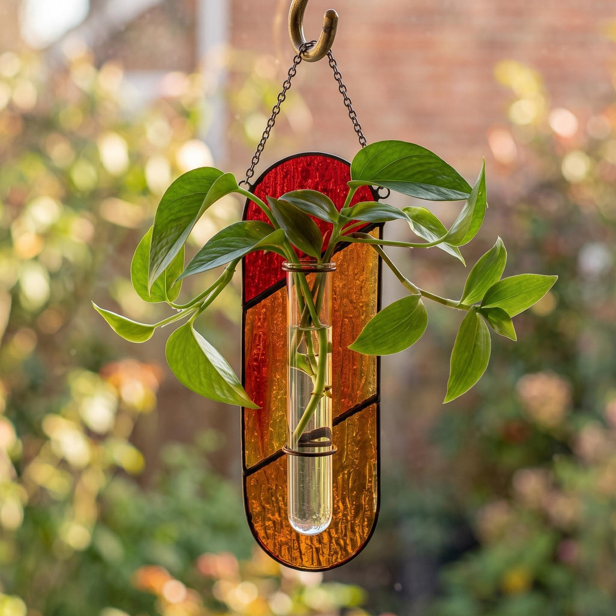 May include: A hanging wall planter featuring a vertical glass test tube held by a stained glass panel with red and orange textured segments. A green leafy plant is rooted in the water-filled tube, which is suspended by a metal chain and hook.