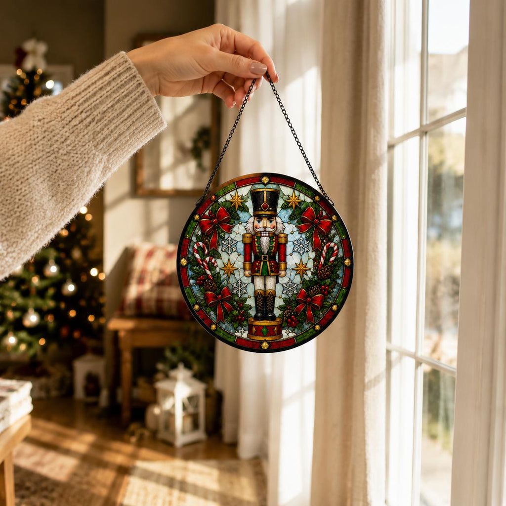 Hand holding a round nutcracker Christmas stained glass suncatcher by its black chain beside a bright holiday window.