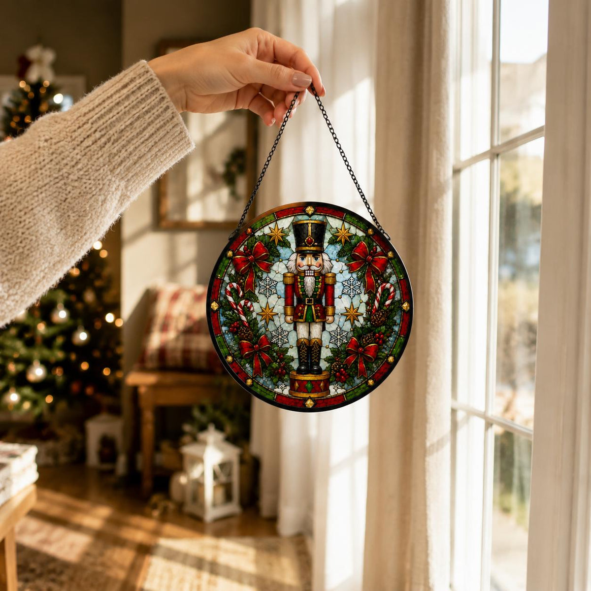 Hand holding a round nutcracker Christmas stained glass suncatcher by its black chain beside a bright holiday window.