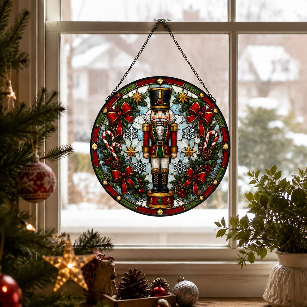 Round nutcracker stained glass window hanging displayed with Christmas greenery, ornaments, pinecones and winter scenery outside.