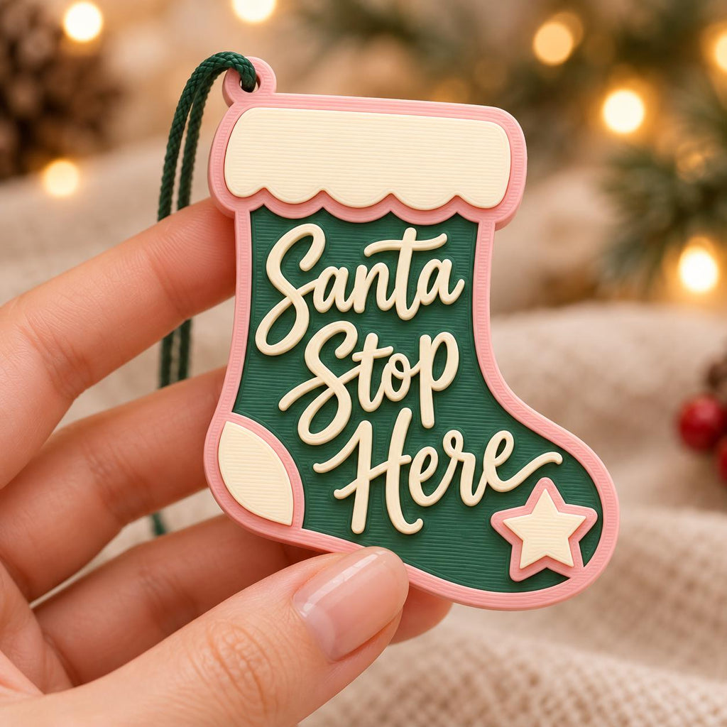May include: A Christmas stocking ornament in dark green with a light pink border and cream-colored accents. The text "Santa Stop Here" is embossed in cream lettering on the front, accompanied by a small cream star at the bottom right corner.