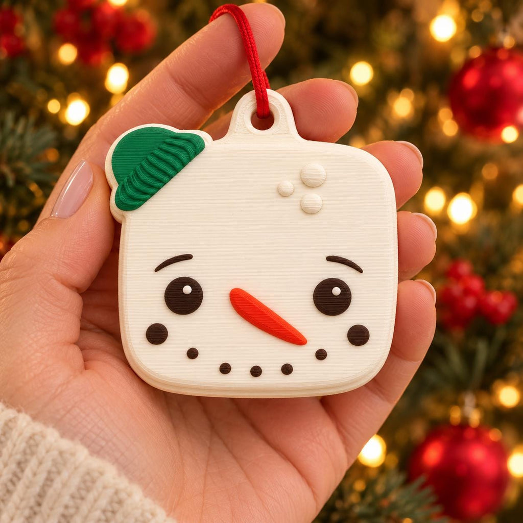 May include: A square-shaped white snowman ornament with rounded corners, featuring a green hat, black eyes, a bright orange carrot nose, and a dotted black mouth. The ornament has a small loop at the top with a red string for hanging.