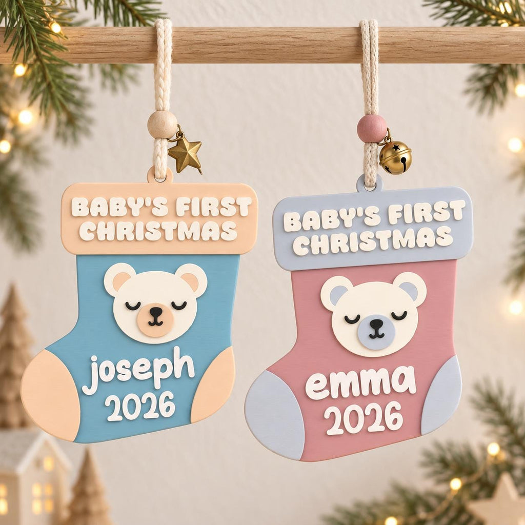 May include: Two stocking-shaped ornaments featuring the text BABY'S FIRST CHRISTMAS. The left ornament is blue with a peach cuff, a bear face, and the name Joseph 2026. The right is pink with a light blue cuff, a bear face, and Emma 2026.