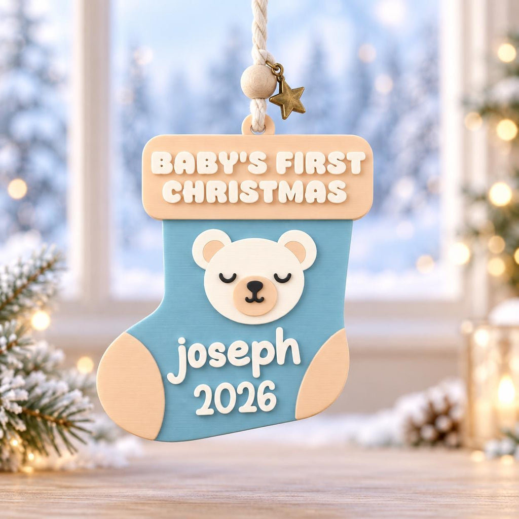 May include: A light blue and cream-colored Christmas stocking ornament featuring a sleeping bear face, the text "BABY'S FIRST CHRISTMAS", the name "joseph", and the year "2026". It hangs from a white rope with a small wooden bead and a bronze star.