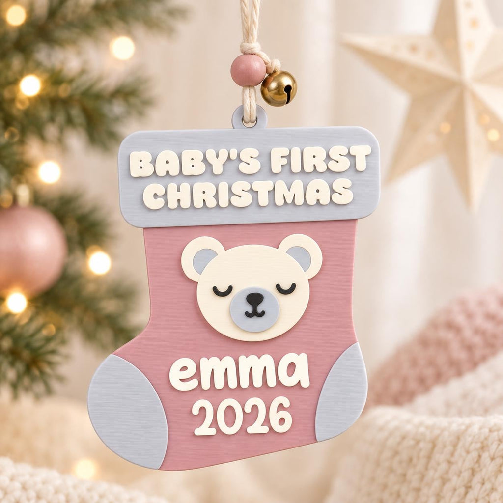 May include: A hanging Christmas stocking ornament in dusty pink and light blue, featuring a cream-colored bear face, the text "BABY'S FIRST CHRISTMAS" at the top, and the name "emma" with the year "2026" printed in white on the front.
