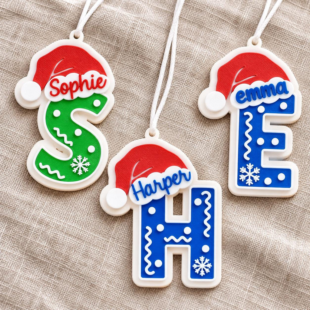 May include: Three letter-shaped Christmas ornaments with white hanging strings. The letters S, H, and E are decorated with red Santa hats, white snowflakes, and wavy lines. The names Sophie, Harper, and emma are printed on the red hat brims.