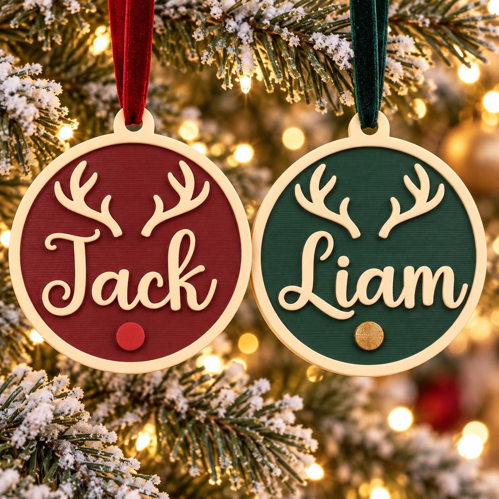 May include: Two circular wooden ornaments with light-colored frames. The left ornament is dark red with the name Jack and cream-colored antlers. The right ornament is dark green with the name Liam and cream-colored antlers. Both have velvet ribbons.