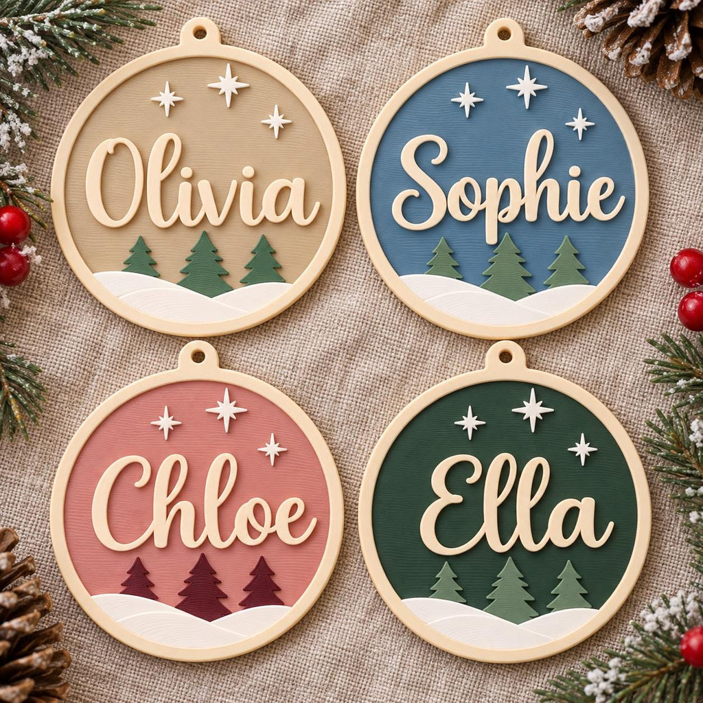 May include: Four circular ornaments with light-colored frames, each featuring a name in raised script lettering: Olivia, Sophie, Chloe, and Ella. The designs include three green or dark red pine trees, white snow-covered ground, and three white stars.