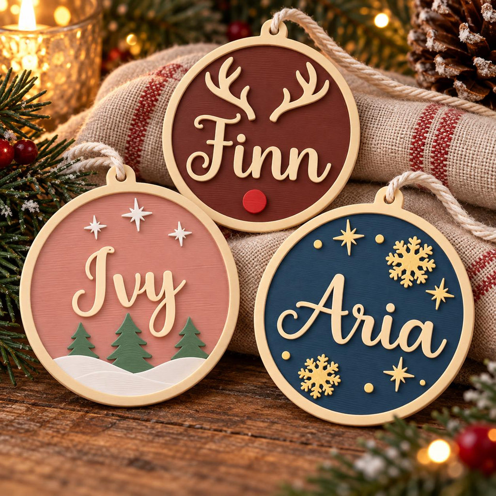 May include: Three circular ornaments with cream-colored borders and hanging loops. One is dark red with cream antlers and the name Finn. One is dusty pink with green trees, white stars, and the name Ivy. One is navy blue with gold snowflakes, stars, and the name Aria.