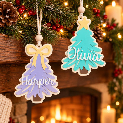 May include: Two layered wooden Christmas tree ornaments hanging by white cords with wooden beads. One tree is light purple with a yellow bow and the name Harper in white script. The other is teal with a yellow star and the name Olivia in white script.