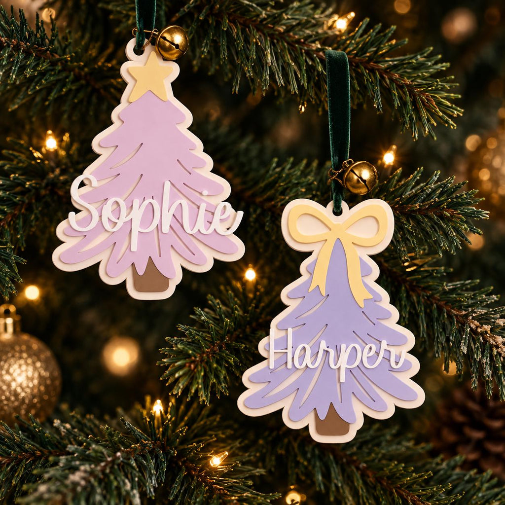 May include: Two layered Christmas tree ornaments made of light purple material with white backing. One features a yellow star and the name Sophie in white script, the other a yellow bow and the name Harper in white script. Both hang from green velvet ribbons with small gold bells.