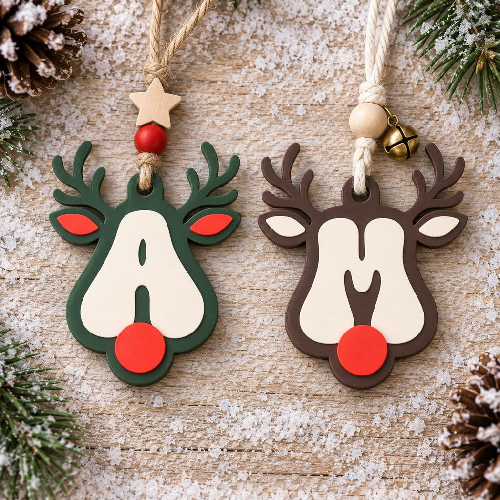 May include: Two reindeer-shaped Christmas ornaments featuring the letters A and M. The left ornament is dark green with a red nose and a star bead. The right ornament is brown with a red nose and a small metal bell. Both hang from twine.