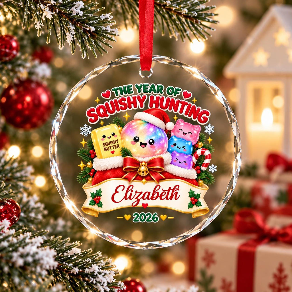 May include: A round, clear glass Christmas ornament hanging from a red ribbon. The design features a rainbow-colored character in a Santa hat, a yellow block labeled SQUISHY BUTTER, and three small cat-like figures. Text reads THE YEAR OF SQUISHY HUNTING, Elizabeth, and 2026.