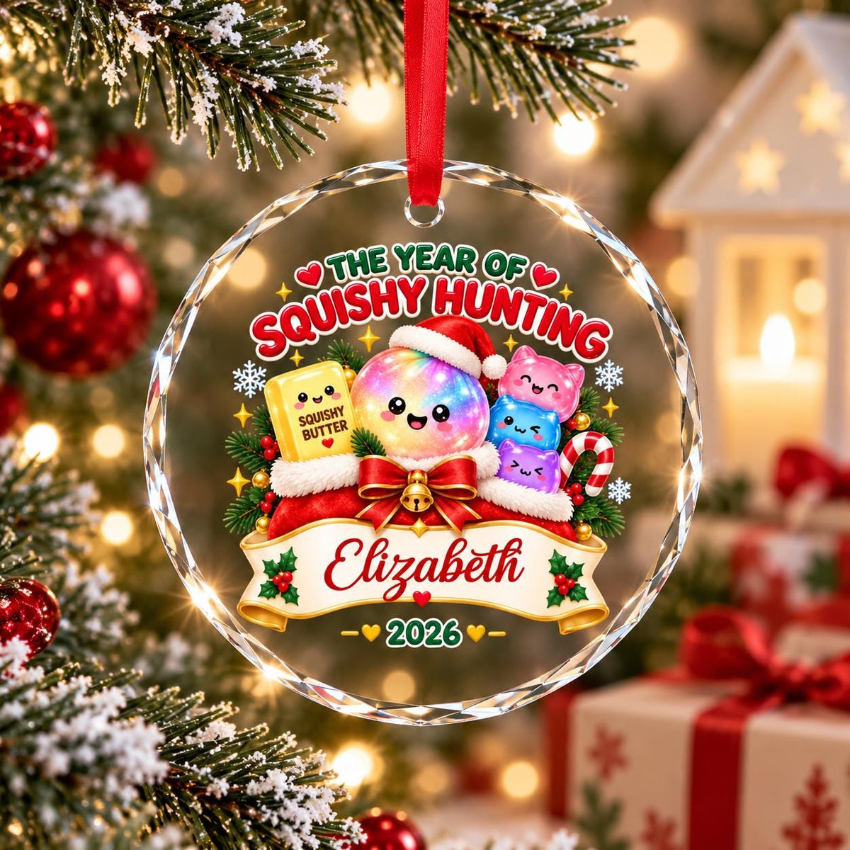 May include: A round, clear glass Christmas ornament hanging from a red ribbon. The design features a rainbow-colored character in a Santa hat, a yellow block labeled SQUISHY BUTTER, and three small cat-like figures. Text reads THE YEAR OF SQUISHY HUNTING, Elizabeth, and 2026.