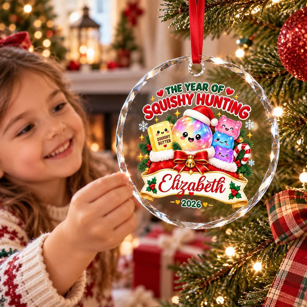 May include: A round, clear glass Christmas ornament featuring a colorful graphic with the text "THE YEAR OF SQUISHY HUNTING", "Elizabeth", and "2026". The design includes a butter stick, a rainbow sphere, and three small pink cat-like figures.