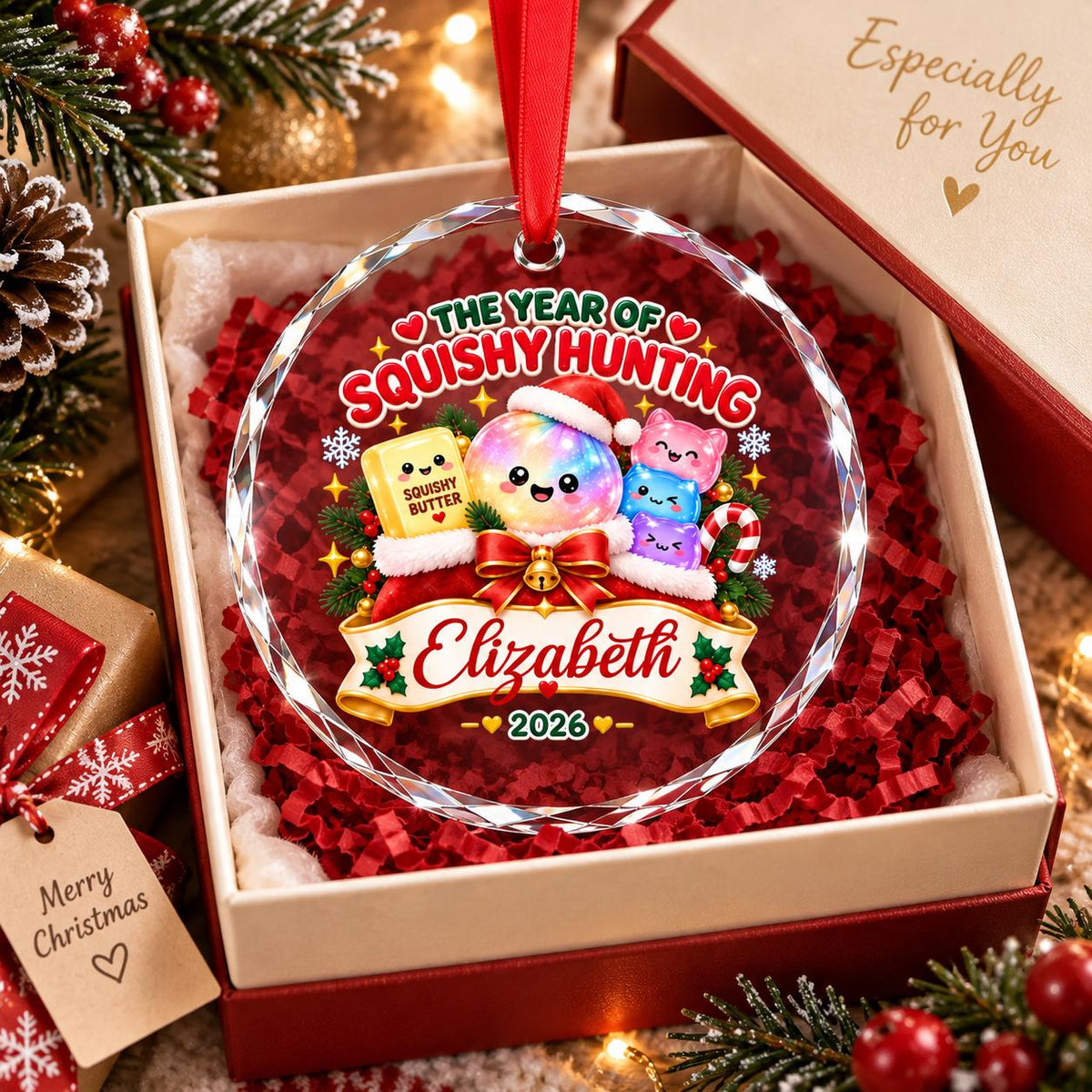 May include: A round, faceted glass Christmas ornament with a red ribbon hanger. The design features a colorful character in a Santa hat, a yellow block labeled SQUISHY BUTTER, and small creatures. Text reads THE YEAR OF SQUISHY HUNTING, Elizabeth, and 2026.