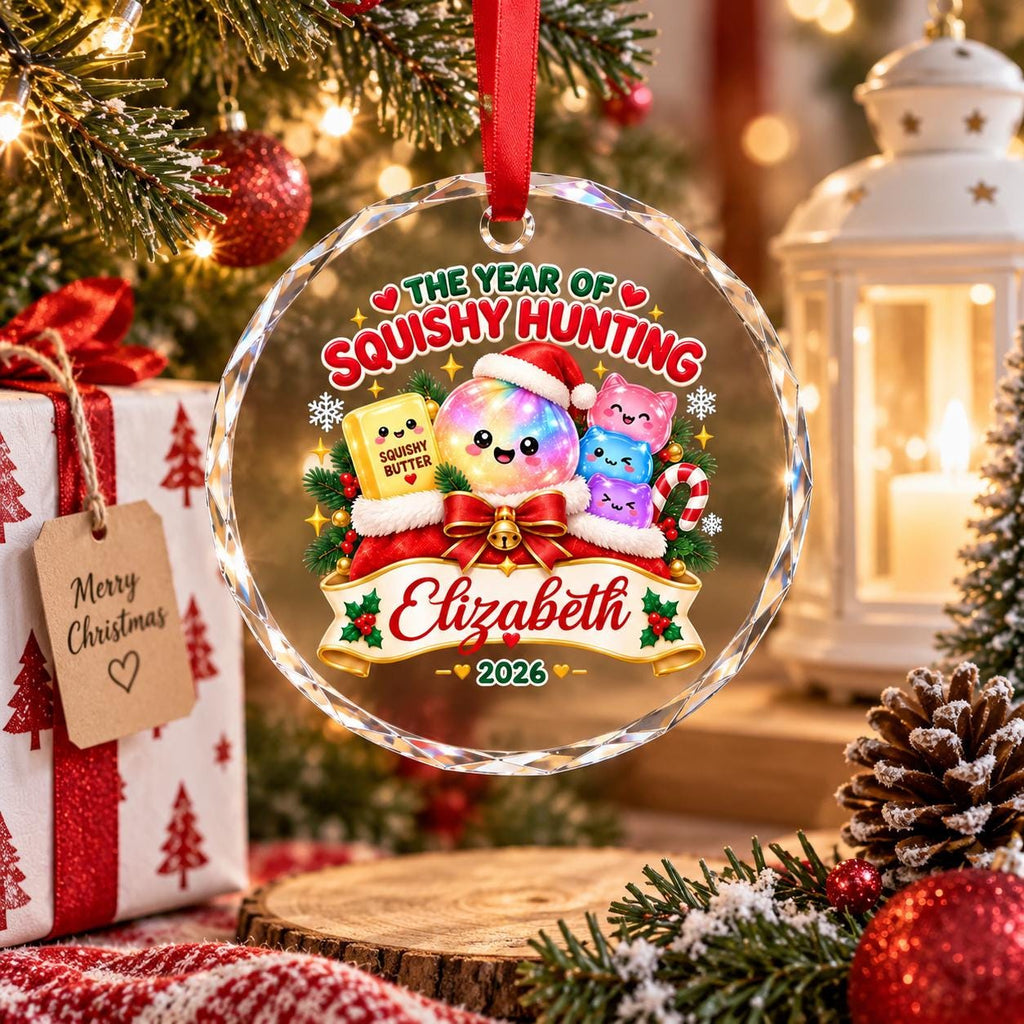 May include: A round, clear glass Christmas ornament featuring a colorful graphic of a squishy butter block, a glowing orb, and stacked squishy toys in Santa hats. The text reads THE YEAR OF SQUISHY HUNTING, Elizabeth, and 2026, with a red ribbon.