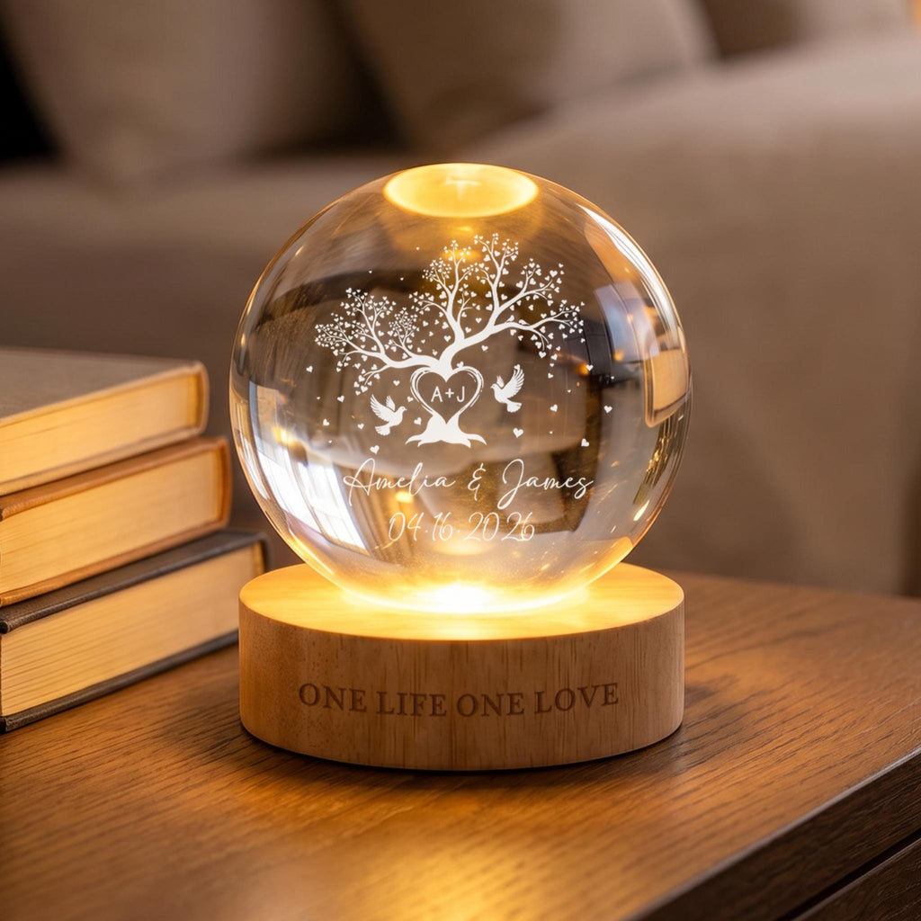 May include: A clear glass sphere lamp featuring an etched tree design with two birds, a heart containing the letters A+J, the names Amelia & James, and the date 04.16.2026. The sphere rests on a light wood base engraved with the text ONE LIFE ONE LOVE.