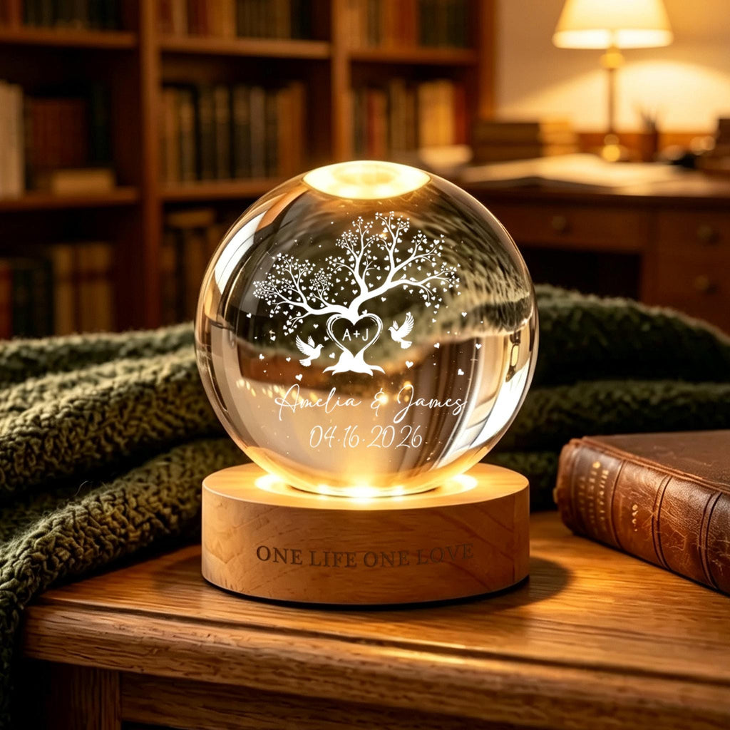 May include: A clear glass sphere lamp resting on a circular wooden base. The sphere features an etched design of a tree, two birds, a heart with the letters A+J, the names Amelia & James, and the date 04.16.2026. The wooden base is engraved with ONE LIFE ONE LOVE.