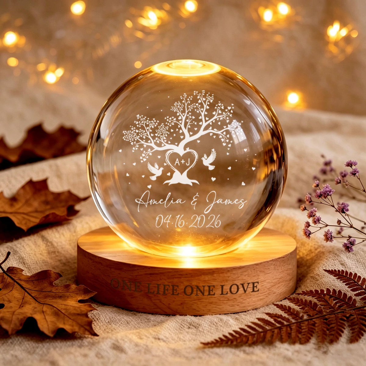 May include: A clear glass sphere lamp on a round wooden base. The sphere features an etched design of a tree with a heart containing the letters A+J, two birds, the names Amelia & James, and the date 04.16.2026. The wooden base is engraved with ONE LIFE ONE LOVE.