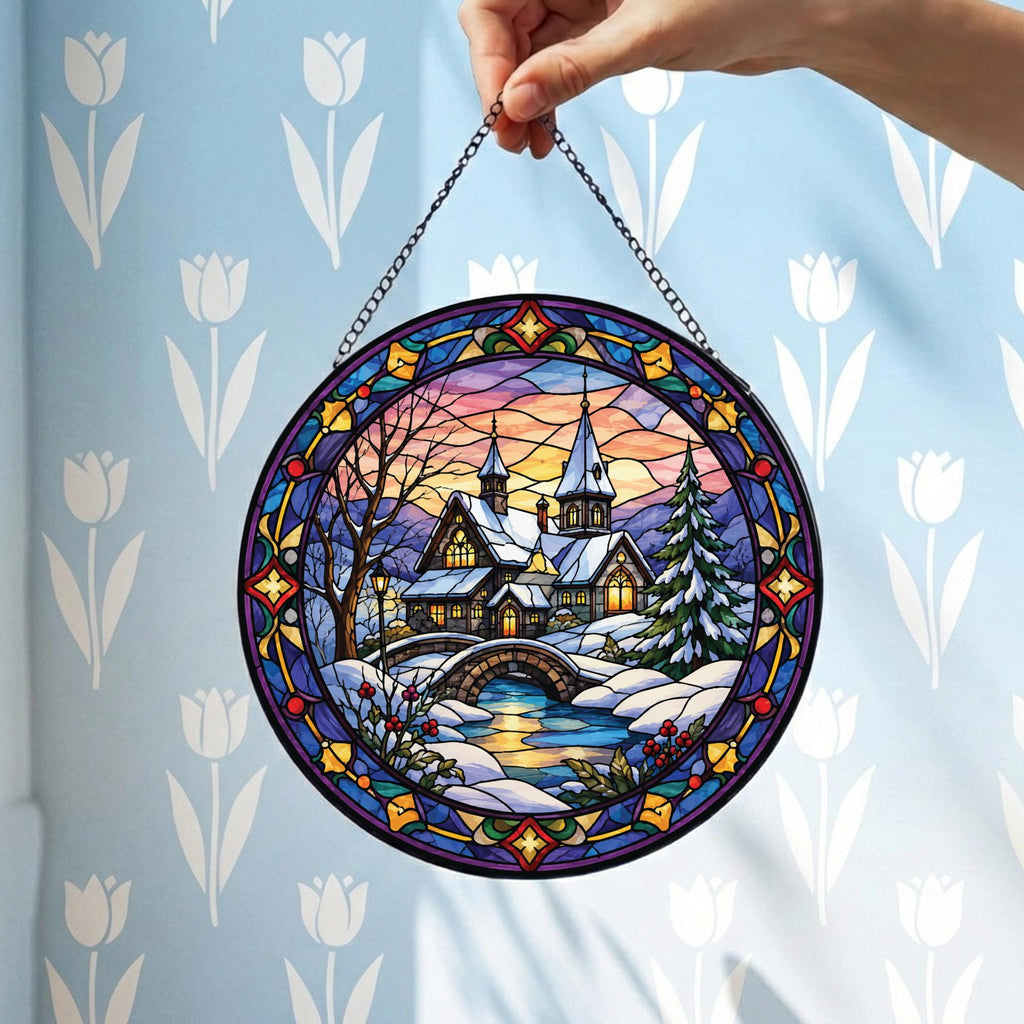 May include: A circular stained glass suncatcher featuring a winter landscape with a snow-covered house, a stone bridge, and a pine tree. The scene is framed by a decorative border of blue, purple, and yellow glass segments and hangs from a metal chain.