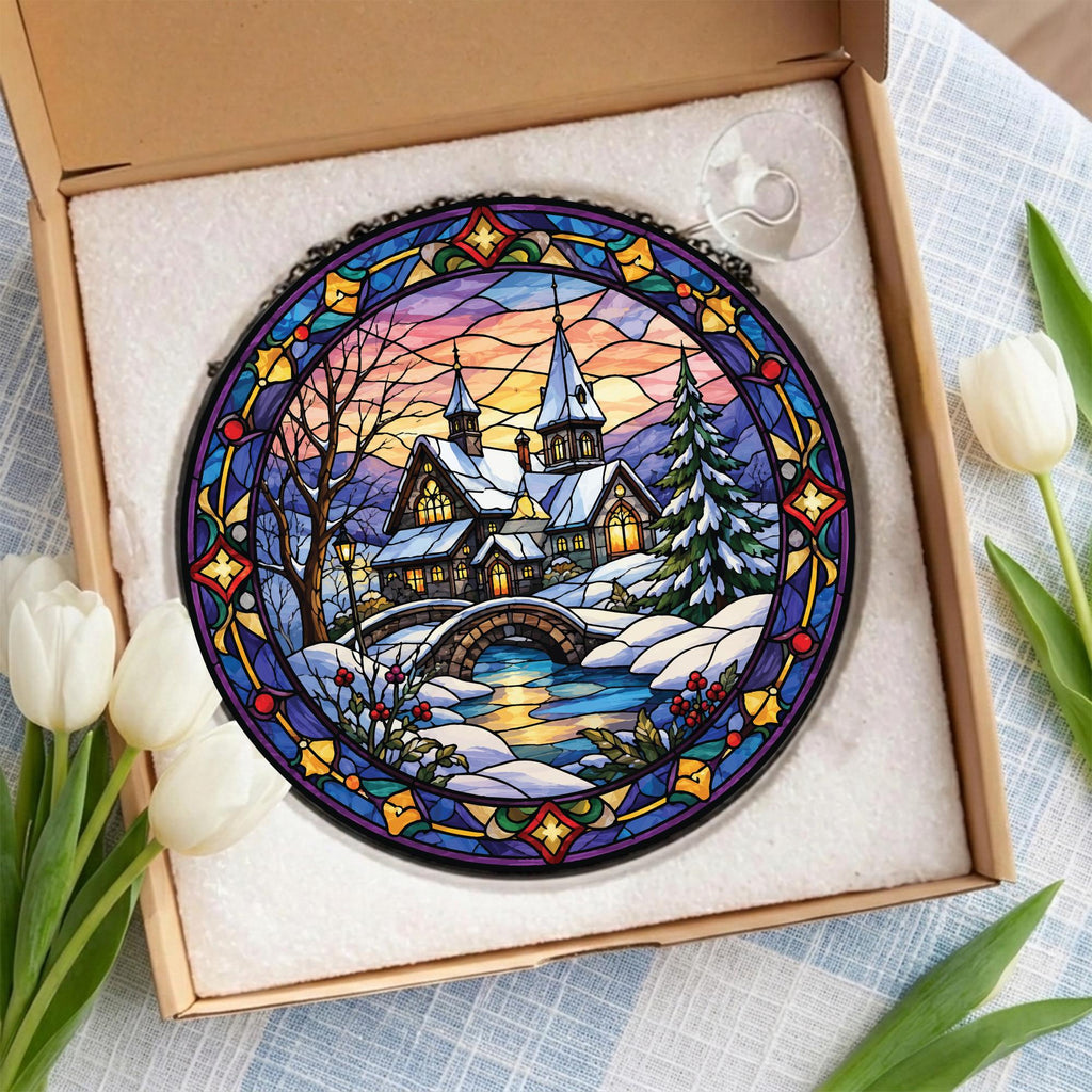 May include: A circular stained glass suncatcher featuring a winter landscape with a snow-covered house, a stone bridge, and a pine tree. The scene is framed by a decorative border of blue, yellow, and red geometric patterns, presented in a box.