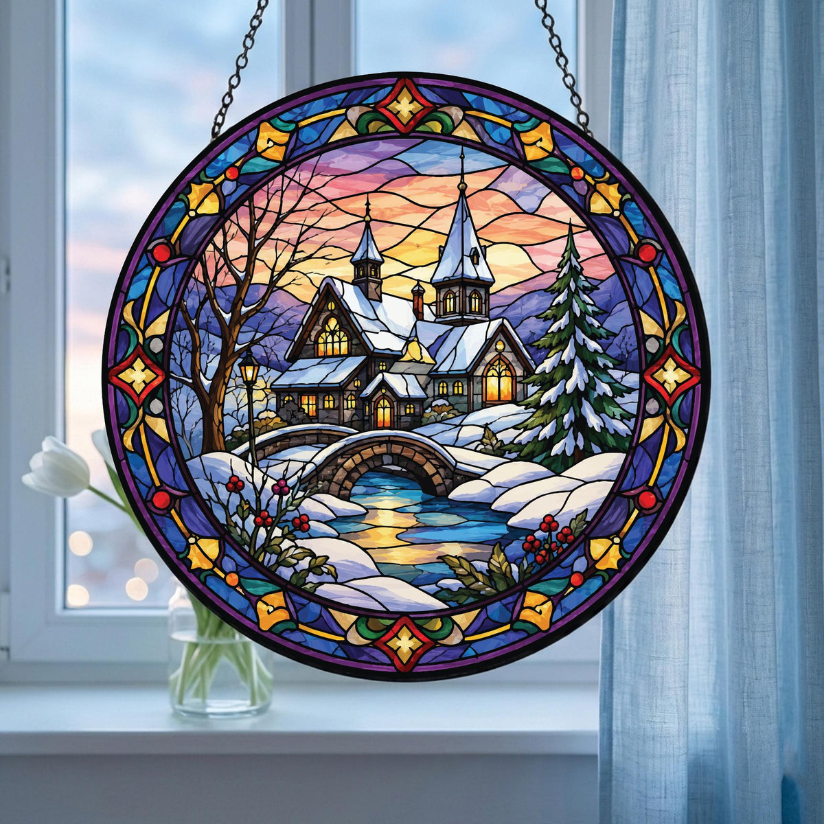 May include: A circular stained glass suncatcher featuring a winter landscape with a snow-covered house, a stone bridge over a stream, and a pine tree. The scene is framed by a decorative border of blue, purple, and yellow glass segments.