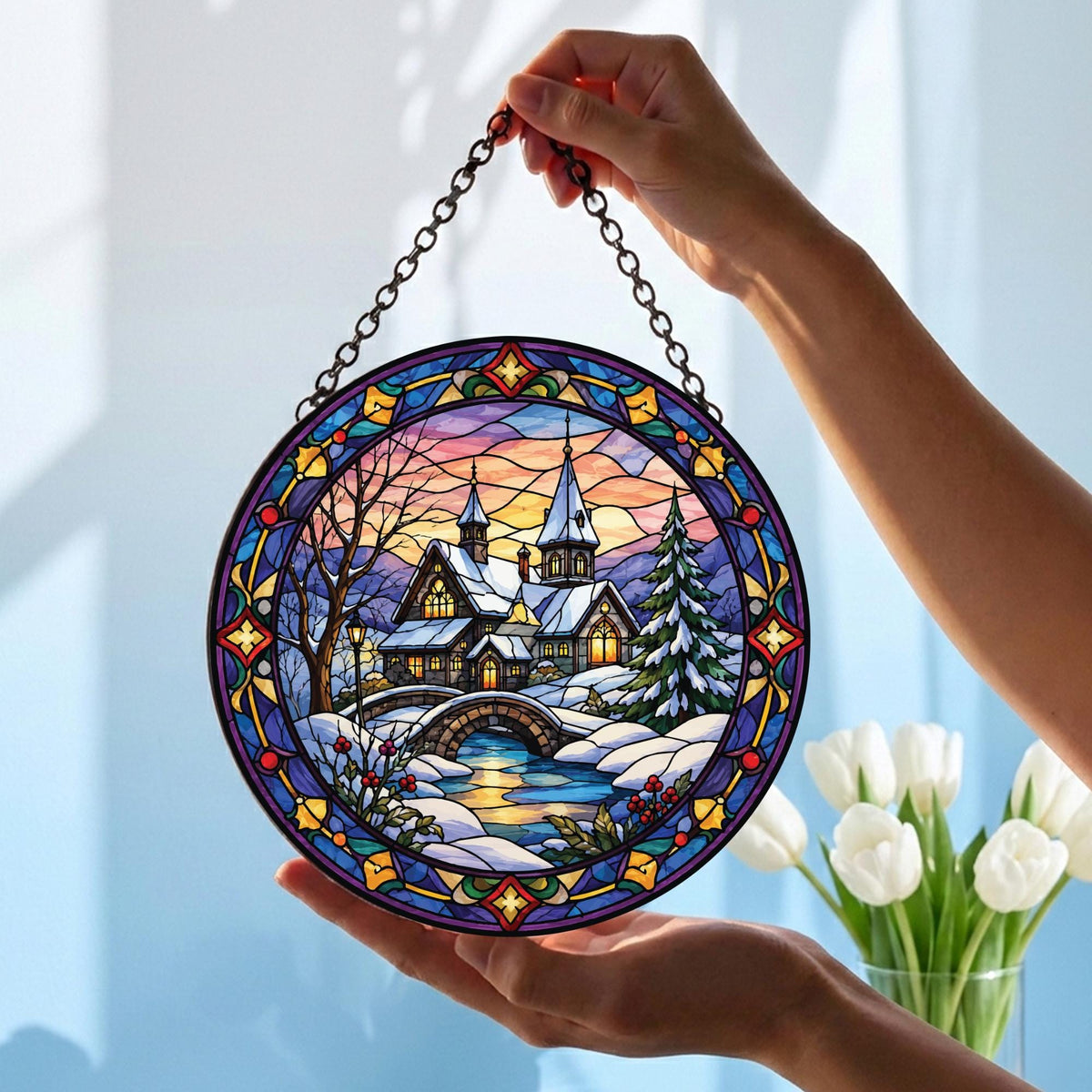 May include: A circular stained glass suncatcher featuring a winter landscape with a snow-covered house, a stone bridge, and a pine tree. The scene is framed by a decorative blue, yellow, and red border and hangs from a metal chain for display.